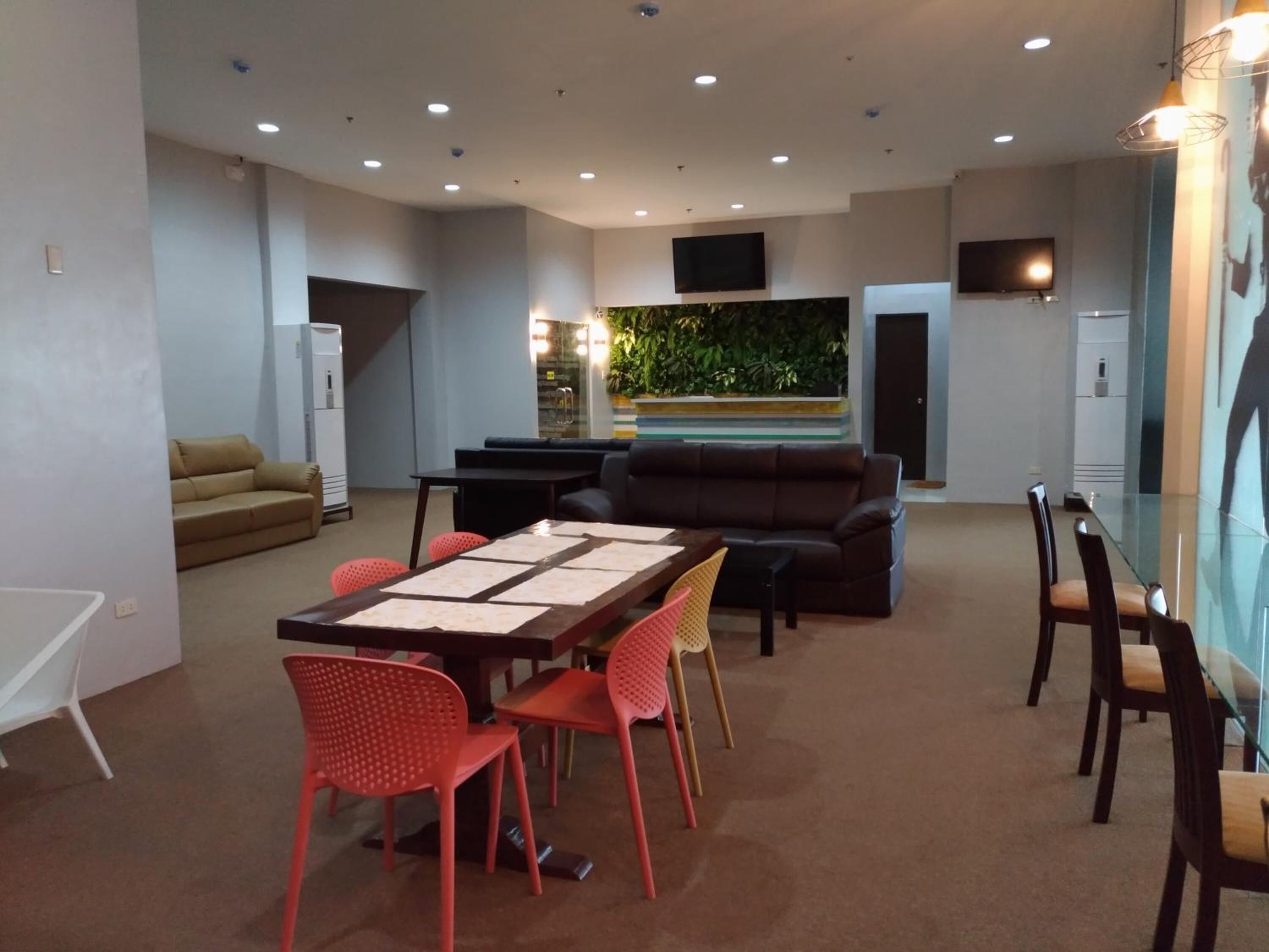 A modern living room with a glass dining table, red chairs, a brown leather sofa, a beige couch, and a small kitchen area with a refrigerator.