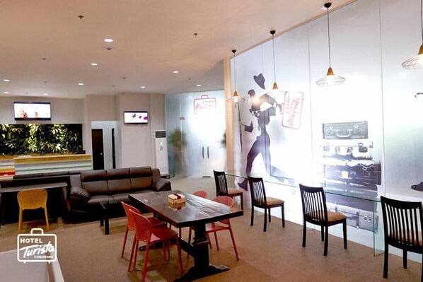 A modern hotel lobby with a dining area, a sofa, and a TV screen.