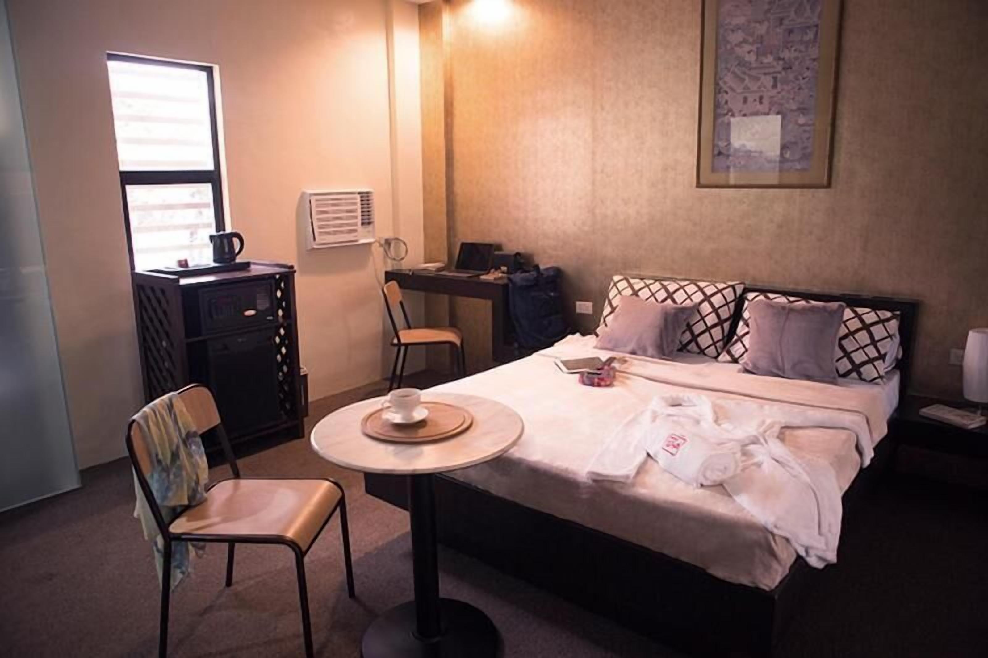 A hotel room with a bed, a desk, a chair, a small table with a cup, and a window.