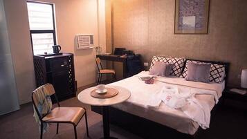 A hotel room with a bed, a desk, a chair, a small table with a cup, and a window.