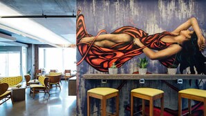 Een moderne bar met een muurschildering van een vrouw in een zwierige jurk, houten barstoelen en een zithoek met gele banken en stoelen.
