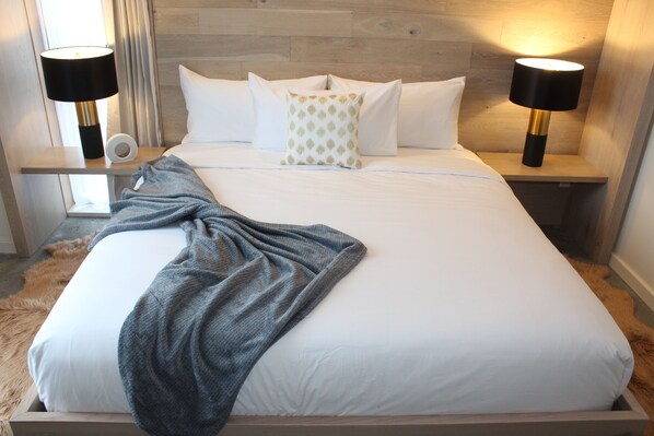 Una cama bien tendida con sábanas blancas, un plaid gris y dos lámparas de noche.