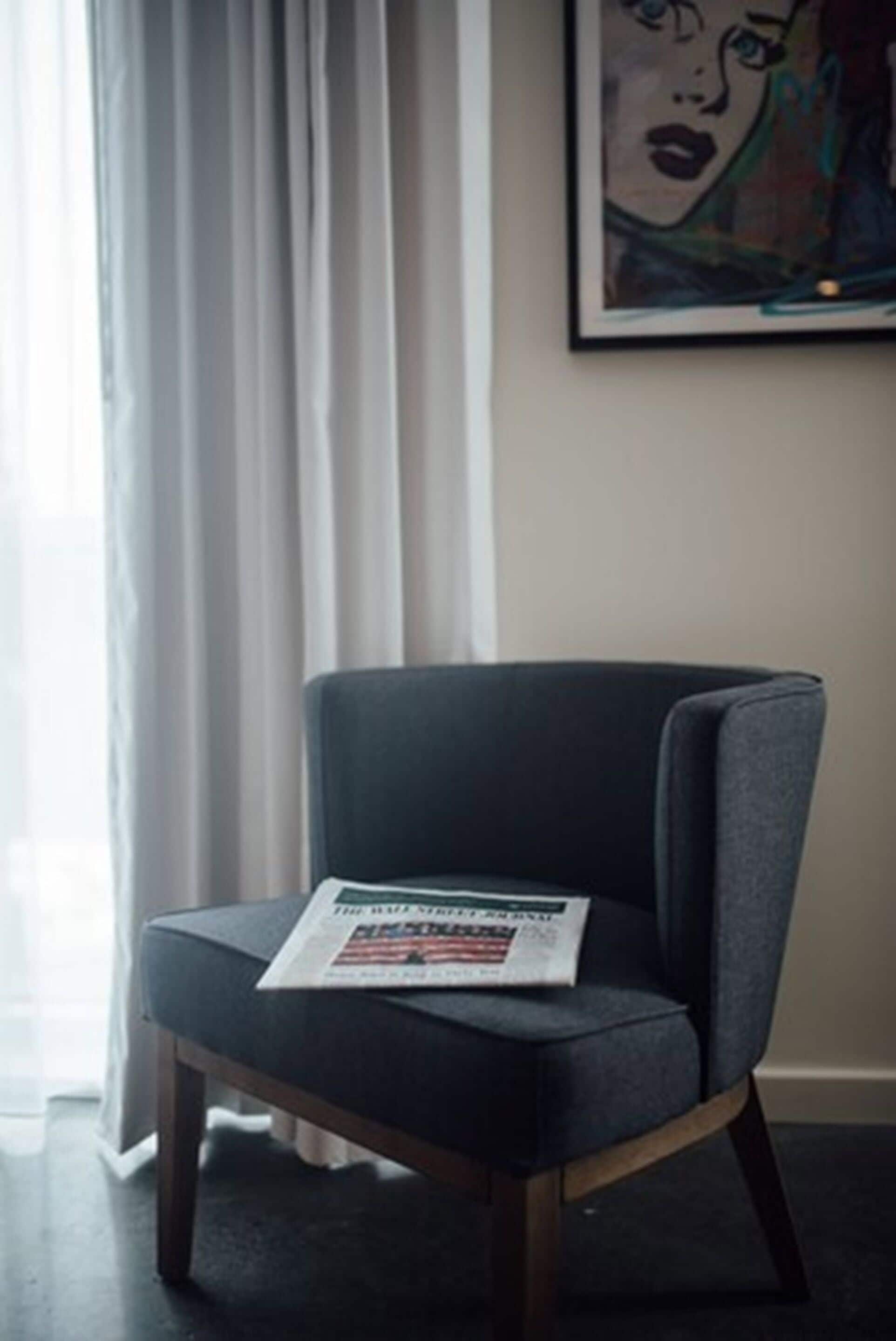Una habitación moderna con un sillón tapizado gris oscuro, un cuadro abstracto enmarcado en la pared y una ventana con cortinas traslúcidas.
