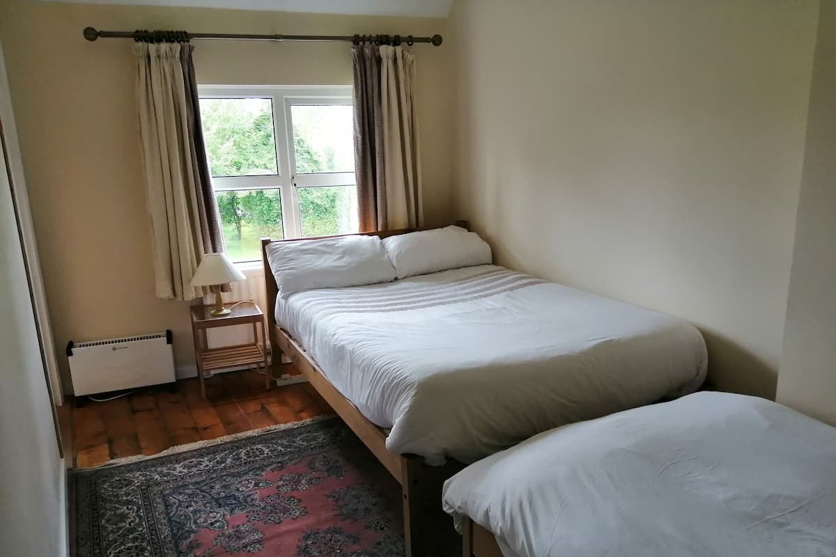 A room with two single beds, a window with curtains, a radiator, a small table with a lamp, and a patterned rug.
