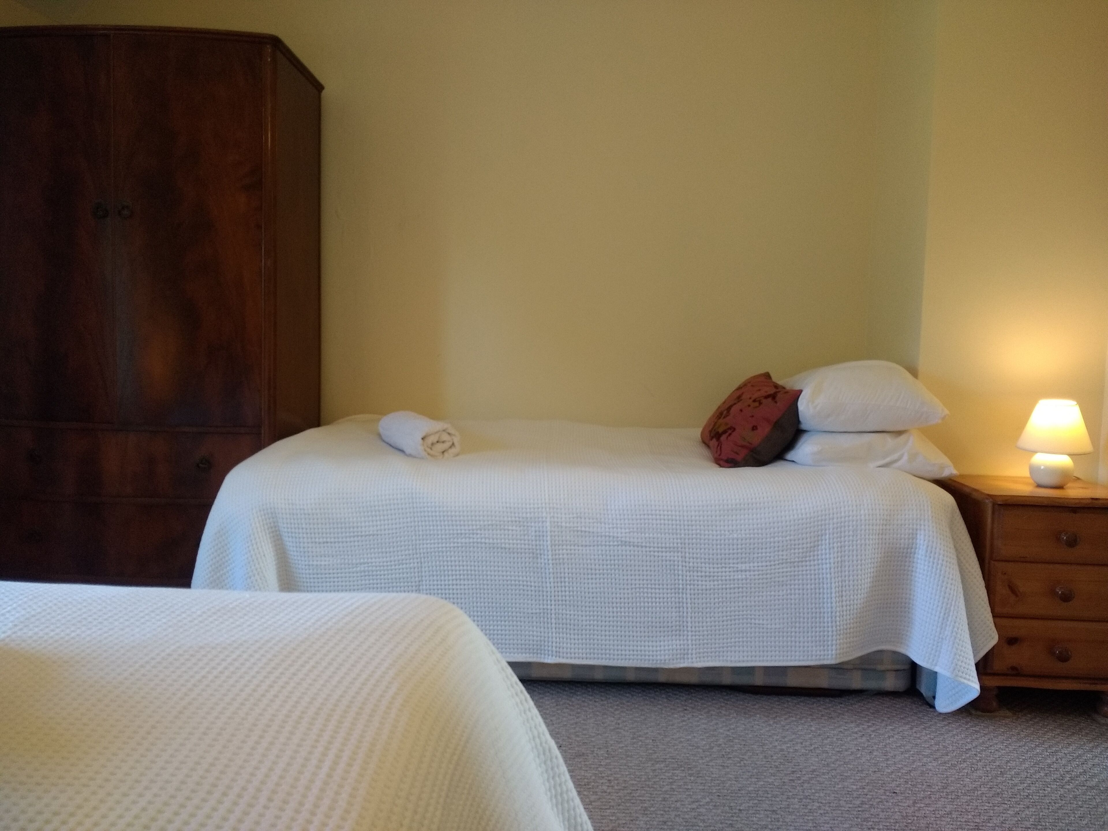 A bedroom with two beds, a wooden wardrobe, a bedside table with a lamp, and a wall-mounted shelf.