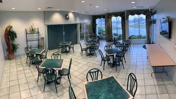 A spacious dining area with multiple tables and chairs, large windows offering a view of the outdoors, and a clock on the wall.