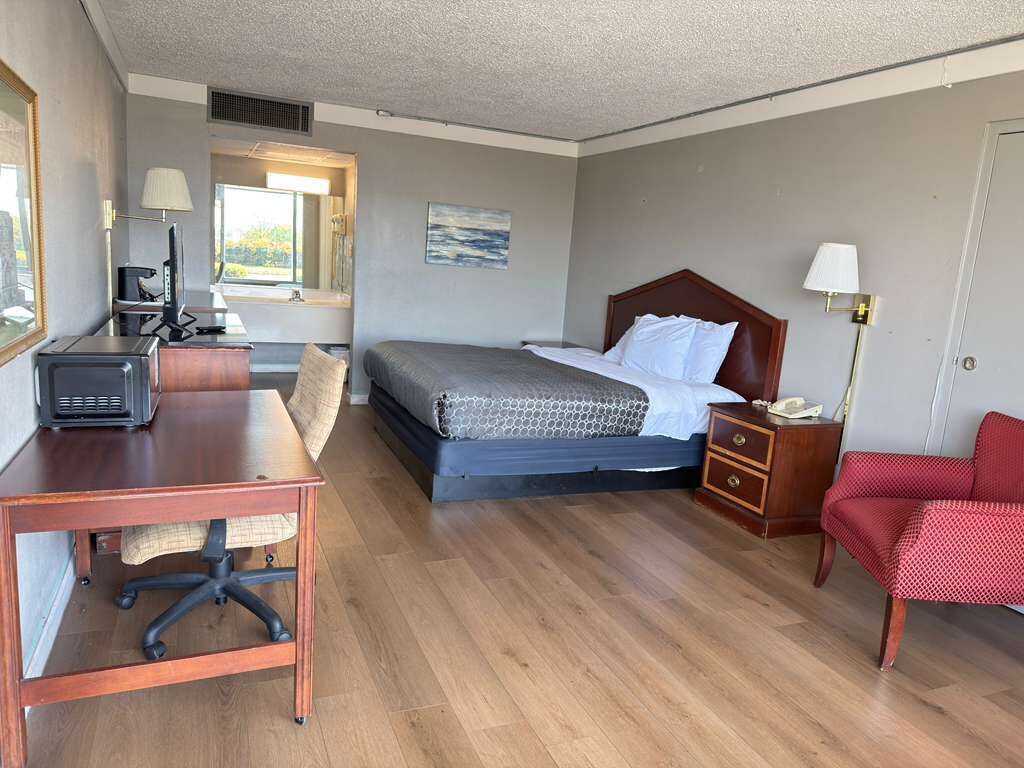 A hotel room with a bed, desk, chair, bedside table, lamp, and a window.