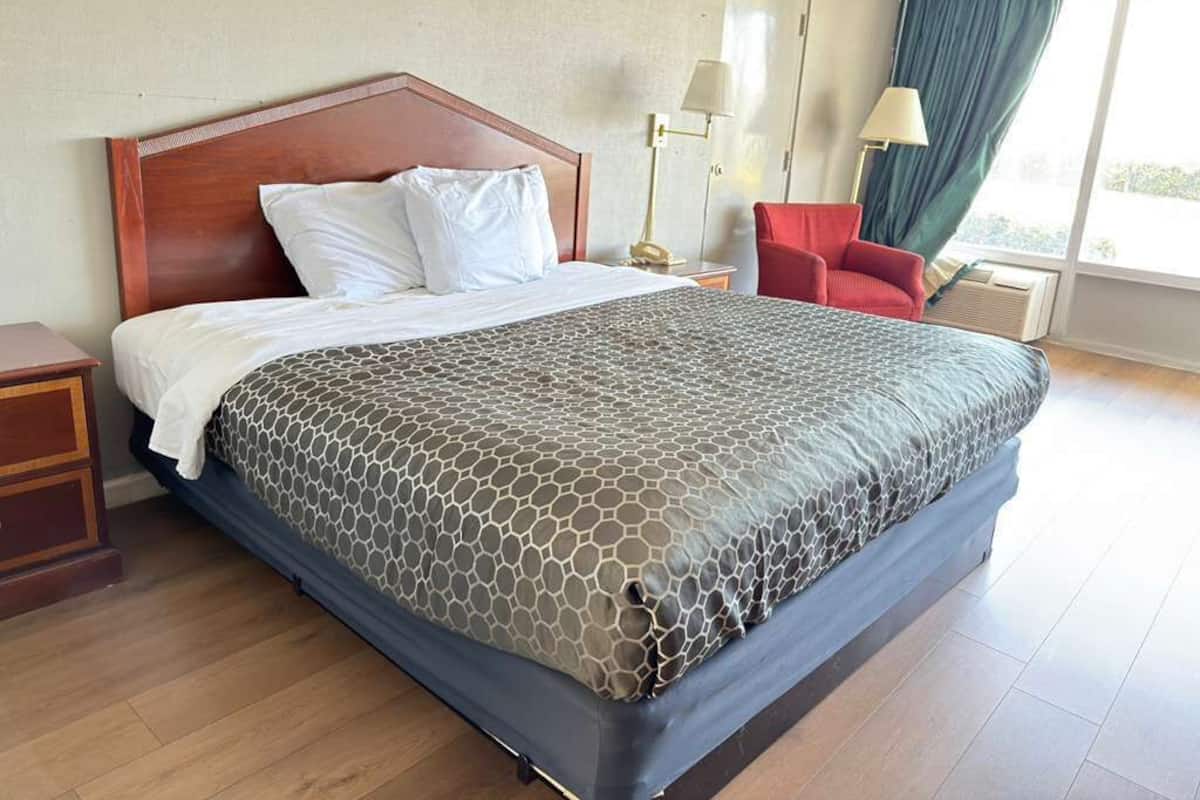 Ein Hotelzimmer mit einem großen Bett, einem Holz-Kopfteil, einem roten Sessel, einem Nachttisch und einem Fenster mit Vorhängen.