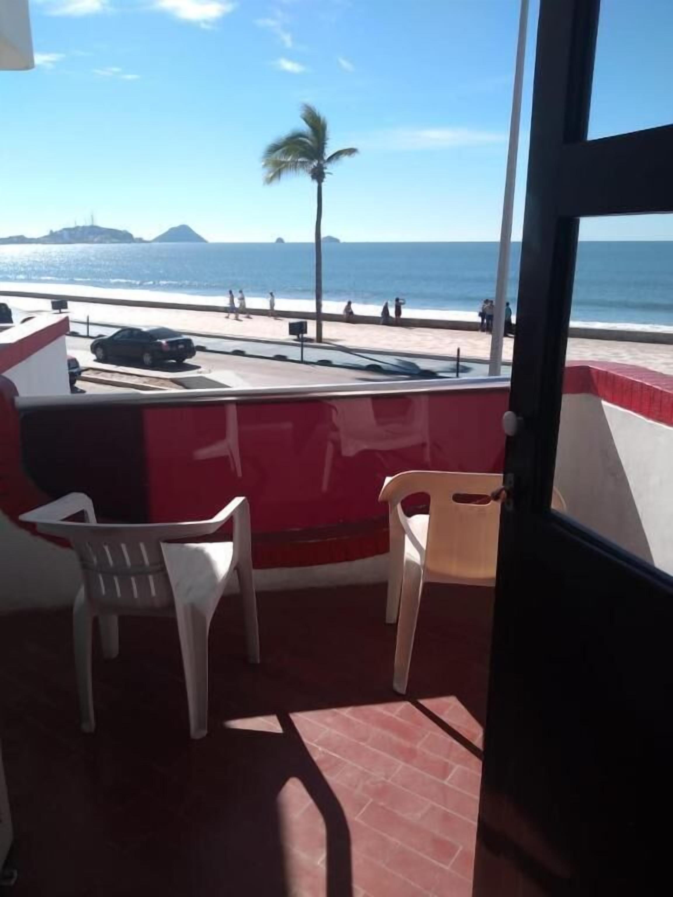Un balcón con vista a la playa y al océano, con dos sillas de plástico blancas y una pared roja y blanca.