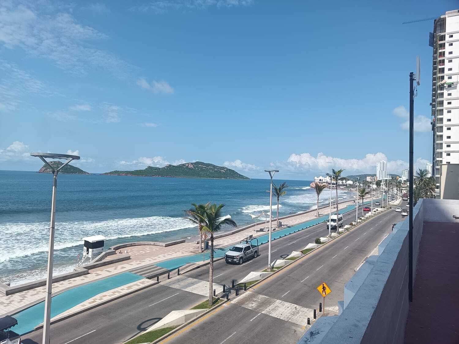 Uma estrada costeira com uma passarela para pedestres, piscina e vista para o oceano e colinas ao longe.