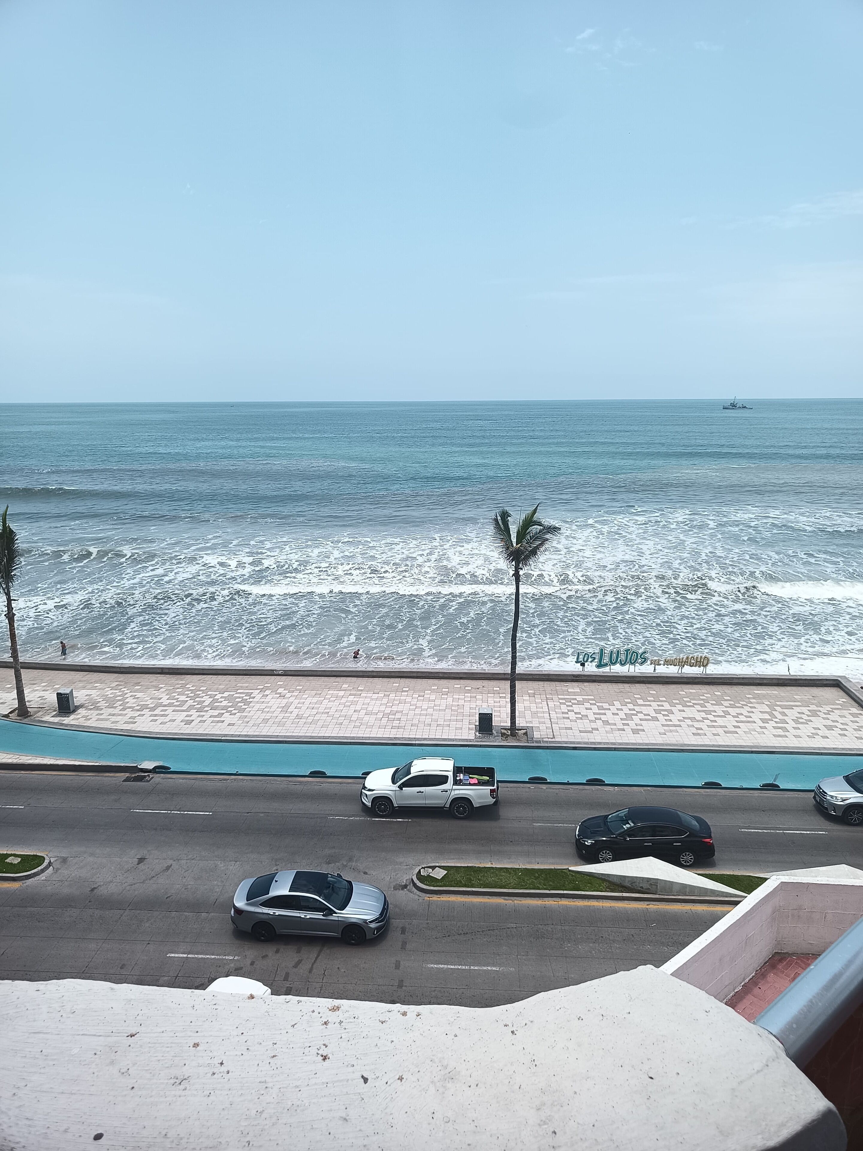 A coastal road with vehicles, a beachside walkway, and a sign reading "Los Lujos del Huchachoco".