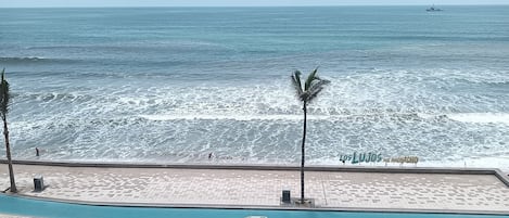 Una carretera costera con vehículos, un paseo marítimo en la playa y un letrero que dice 'Los Lujos del Huchachoco'.