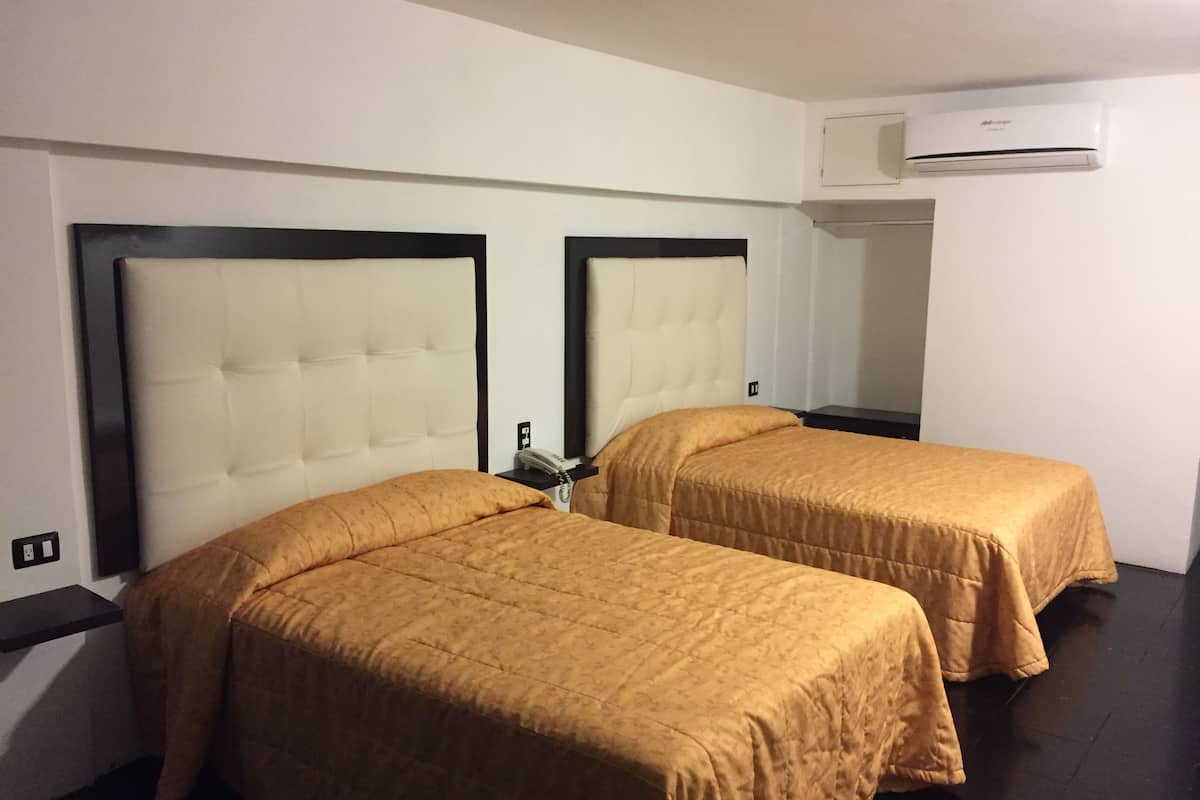 A hotel room with two beds, each with a headboard and a bedside table. There is an air conditioning unit mounted on the wall above the beds.