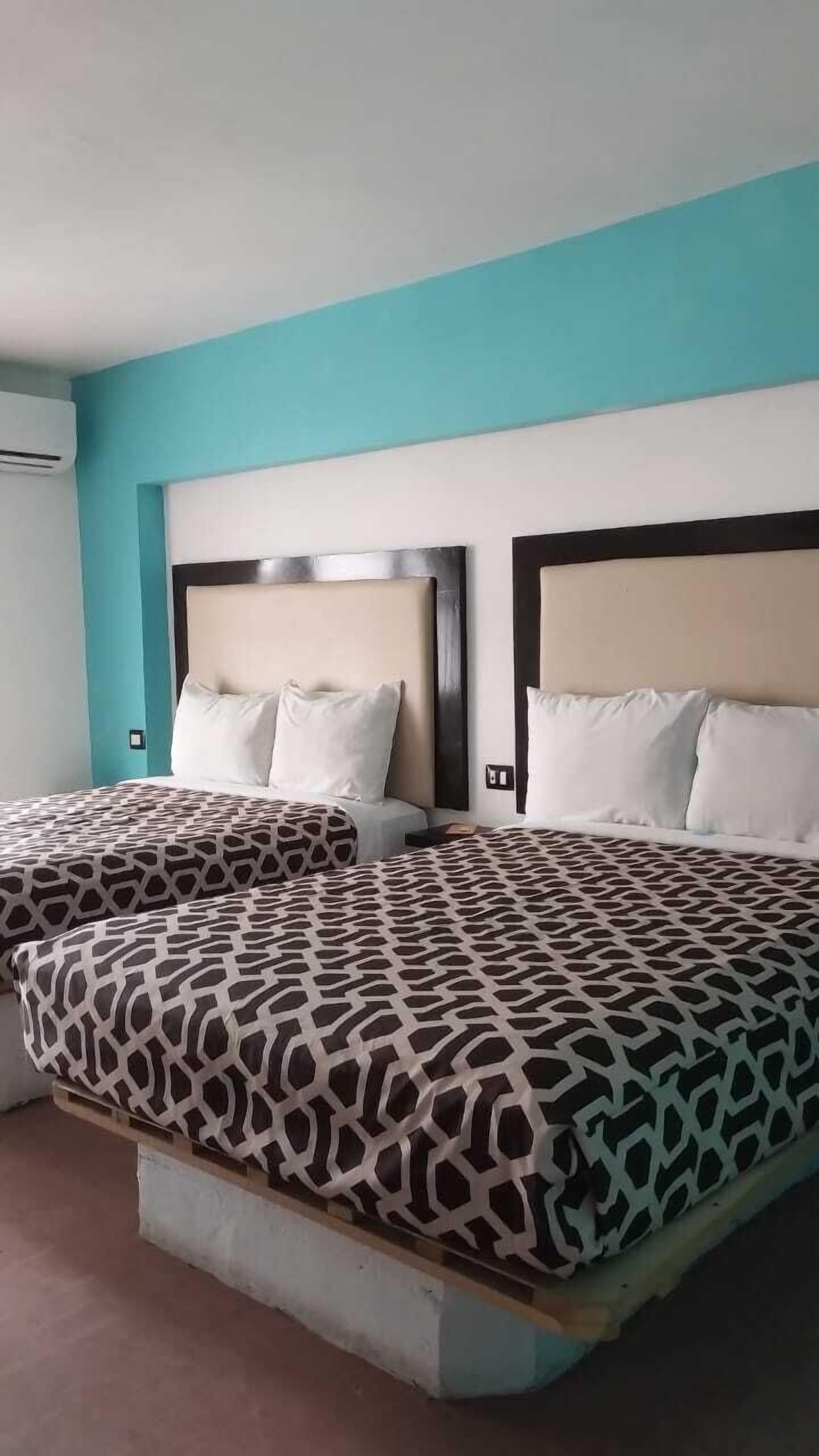 Une chambre d’hôtel avec deux lits, chacun recouvert d’une couvre-lit à motifs et agrémenté d’oreillers blancs. Les murs de la chambre sont de couleur turquoise et le lit est doté d’une tête de lit en bois.