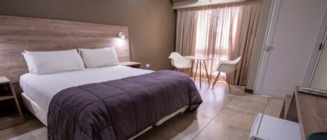 A hotel room with a bed, bedside tables, a desk with a chair, a small refrigerator, and a window with curtains.
