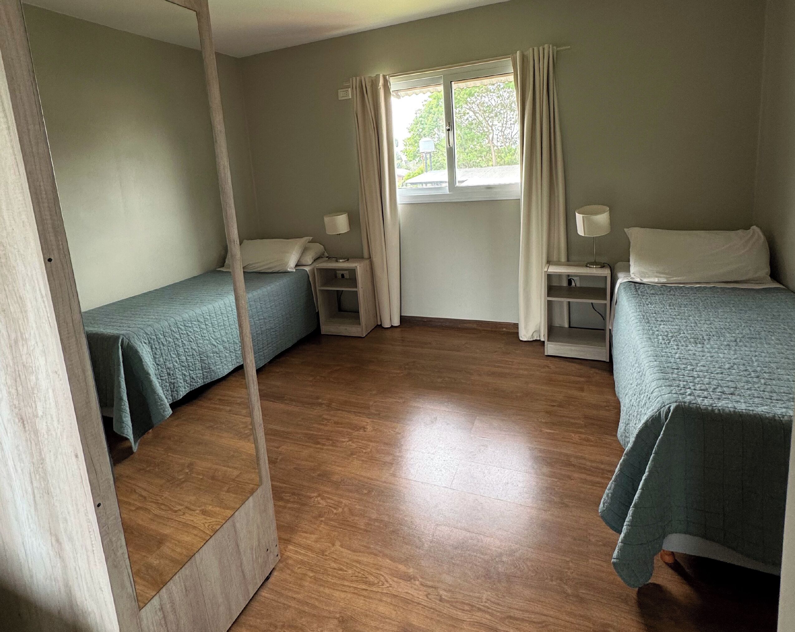 Una habitación con dos camas individuales, suelo de madera, un ventanal con cortinas y mesitas de noche con lámparas.