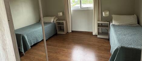 Una habitación con dos camas individuales, suelo de madera, un ventanal con cortinas y mesitas de noche con lámparas.