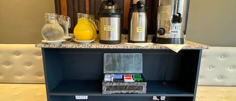 Una encimera de cocina con diversos electrodomésticos y recipientes, incluyendo una cafetera, un dispensador de agua caliente y un termo. Debajo de la encimera, hay un estante con una caja de pañuelos.