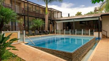 Una piscina rectangular con borde de piedra, rodeada por un patio con sillas de descanso y un área de estar techada.