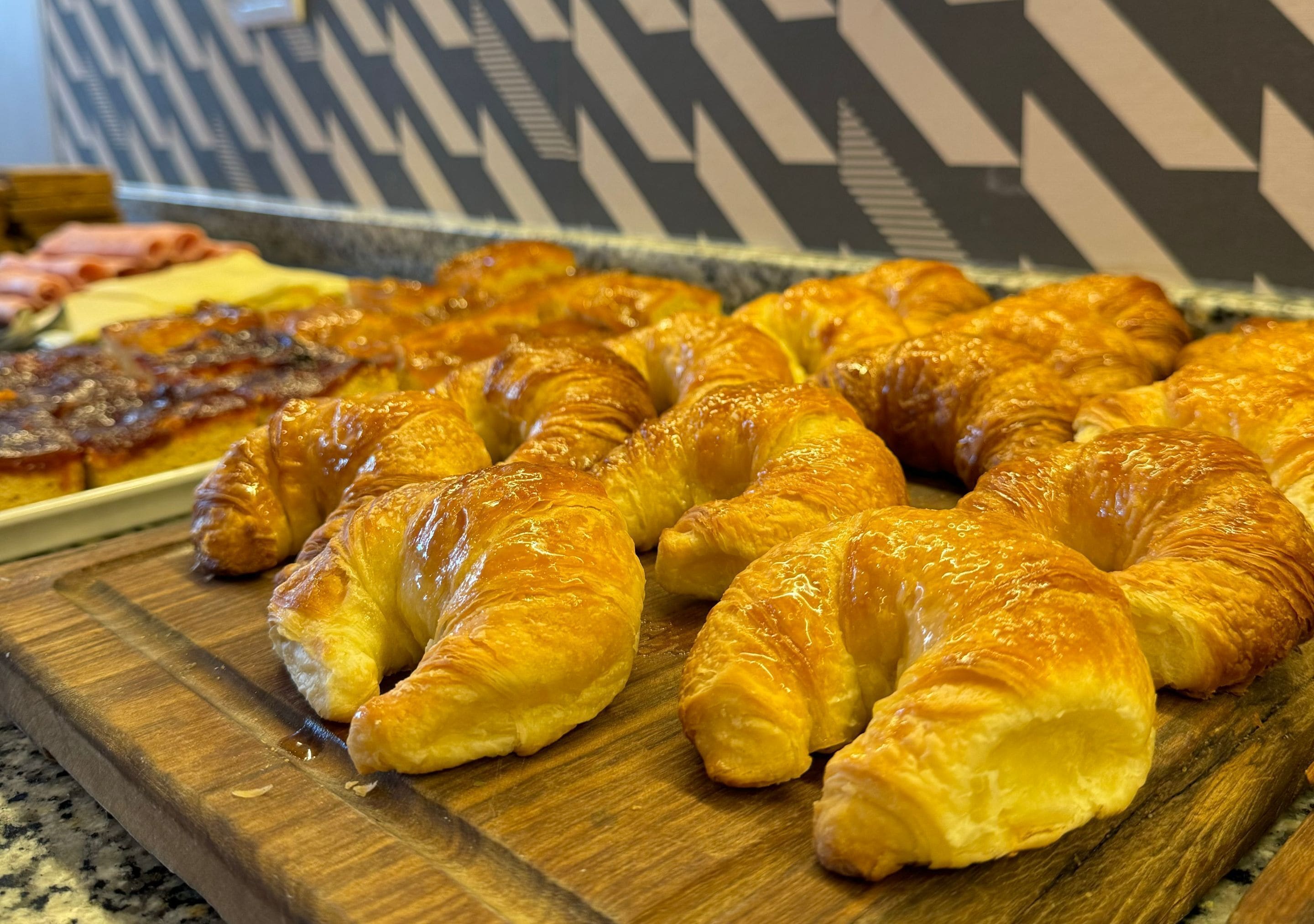 A wooden board with several golden-brown croissants and a tray of sliced meats.
