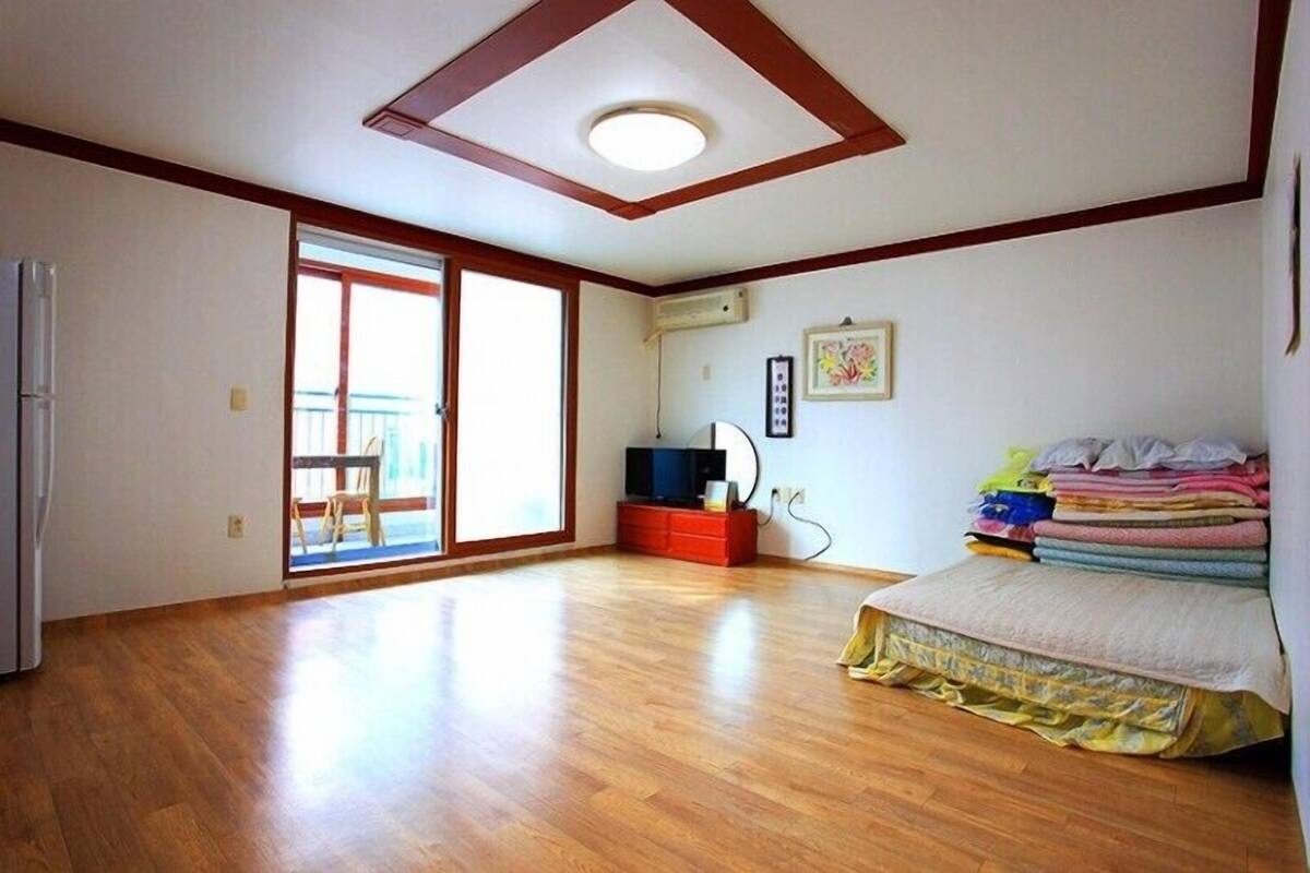 Ein geräumiges Zimmer mit Holzboden, einem Bett mit farbenfroher Bettwäsche, einem kleinen Fernseher und einer Schiebetür zum Balkon.