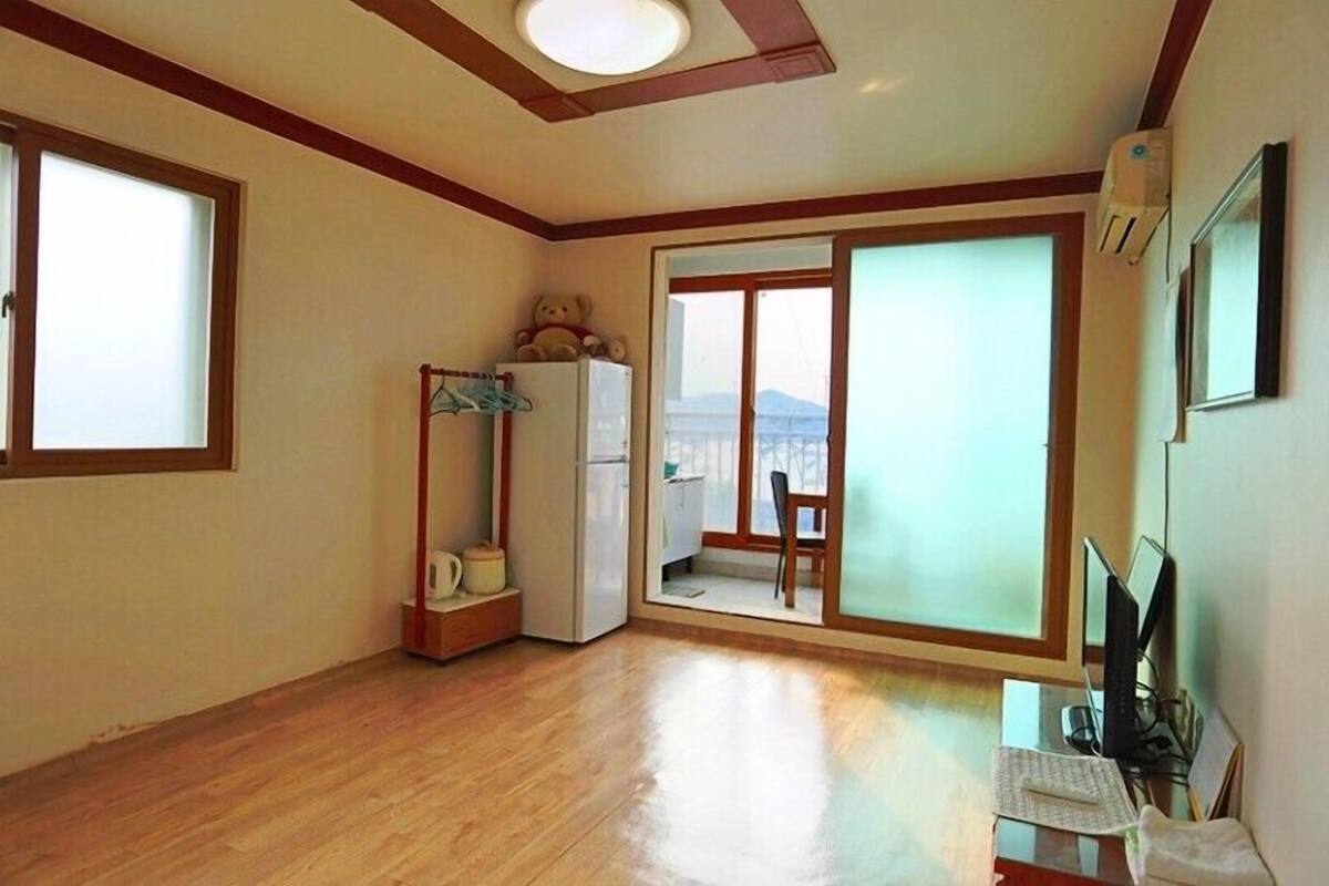 Ein geräumiges Zimmer mit Holzboden, einer großen Schiebetür zum Balkon, einem weißen Kühlschrank und einem kleinen Tisch mit Stuhl.