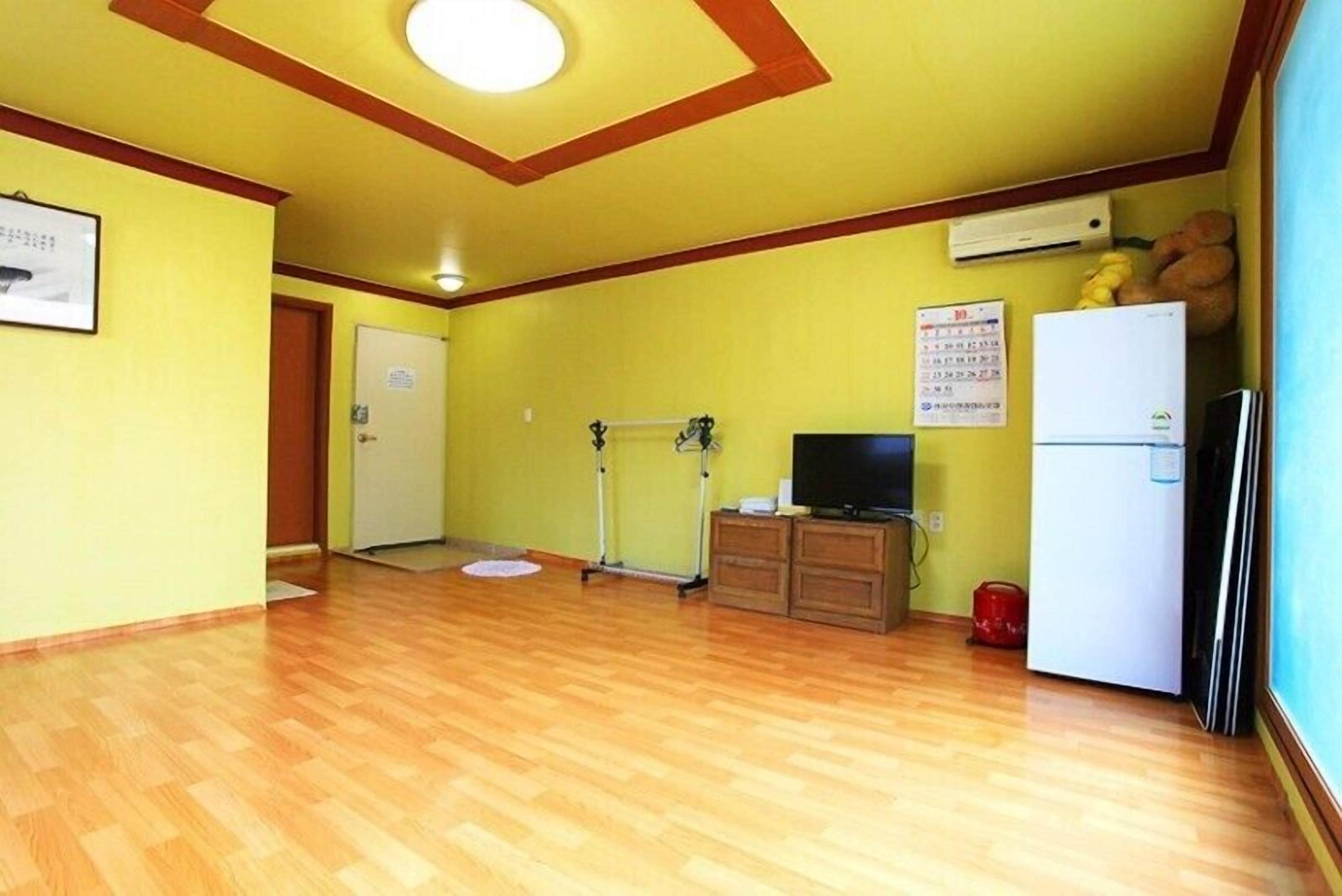 A room with wooden flooring, a television, a refrigerator, and a door leading to another room.