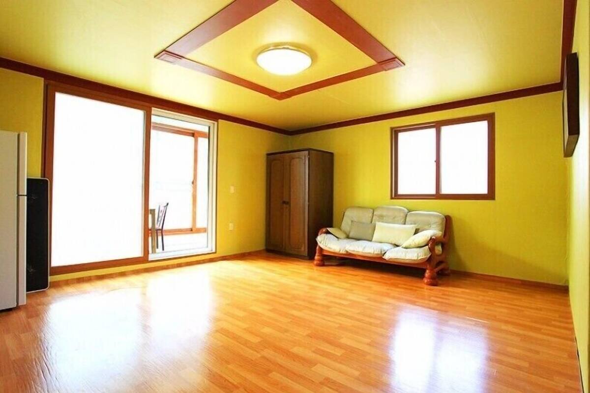 Ein geräumiger Raum mit Holzboden, einer Couch, einem Schrank und großen Fenstern, die viel Tageslicht hereinlassen.
