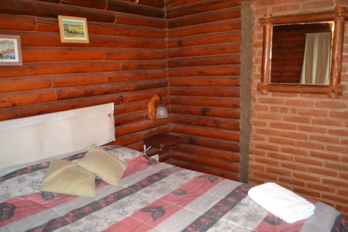 Una habitación tipo cabaña de madera con una cama, un estante pequeño y una ventana con cortina.