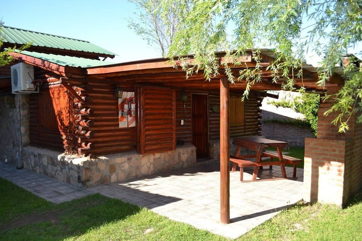 Una cabaña rústica de troncos con un porche cubierto, base de piedra y una mesa y banco de madera.