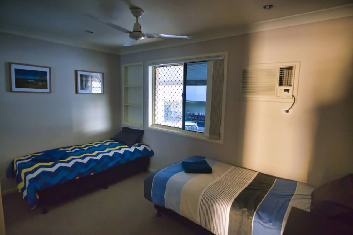 A bedroom with two single beds, a ceiling fan, a window with blinds, and a wall-mounted air conditioning unit.