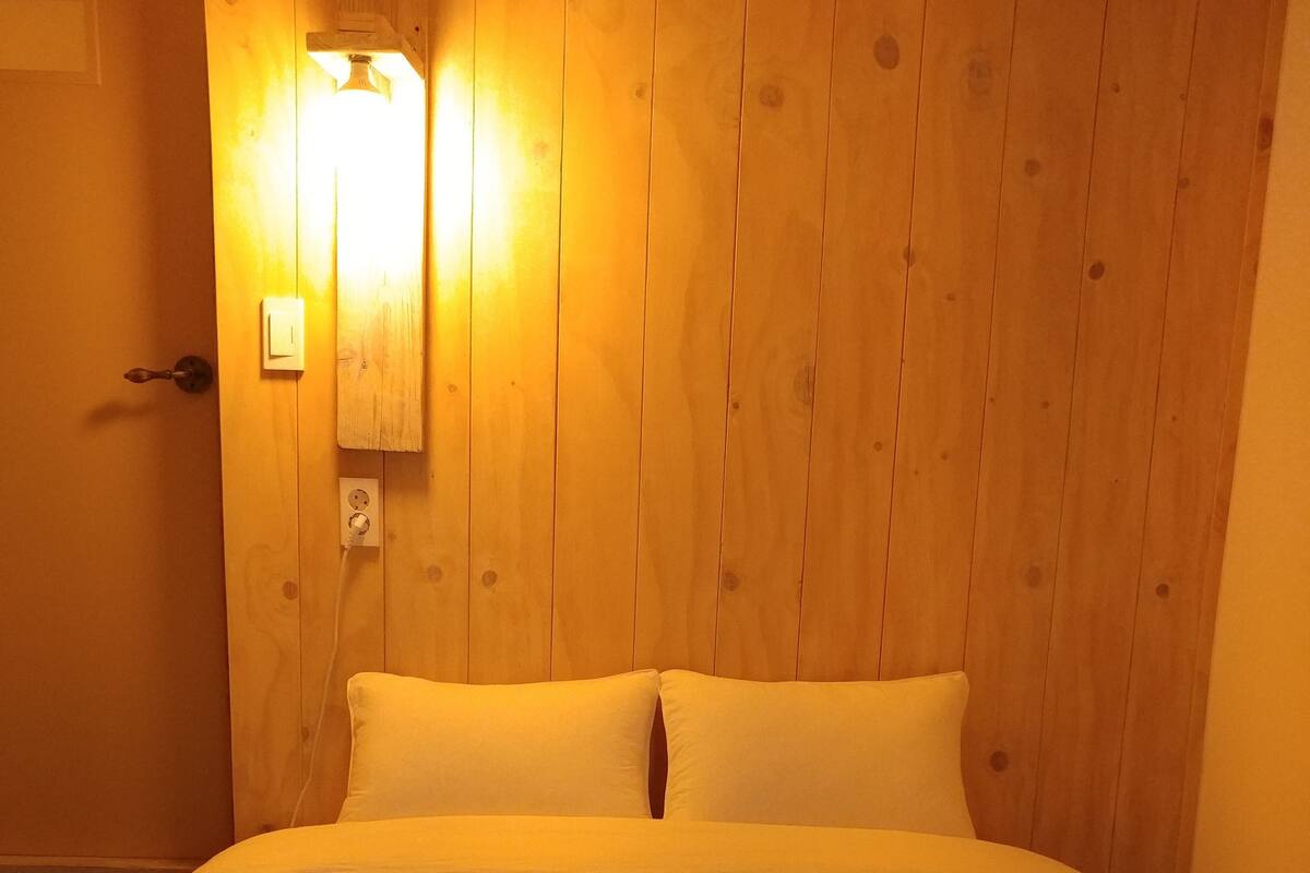 Una habitación con pared de madera, una cama con ropa blanca, una lámpara de pared y una puerta con pomo.