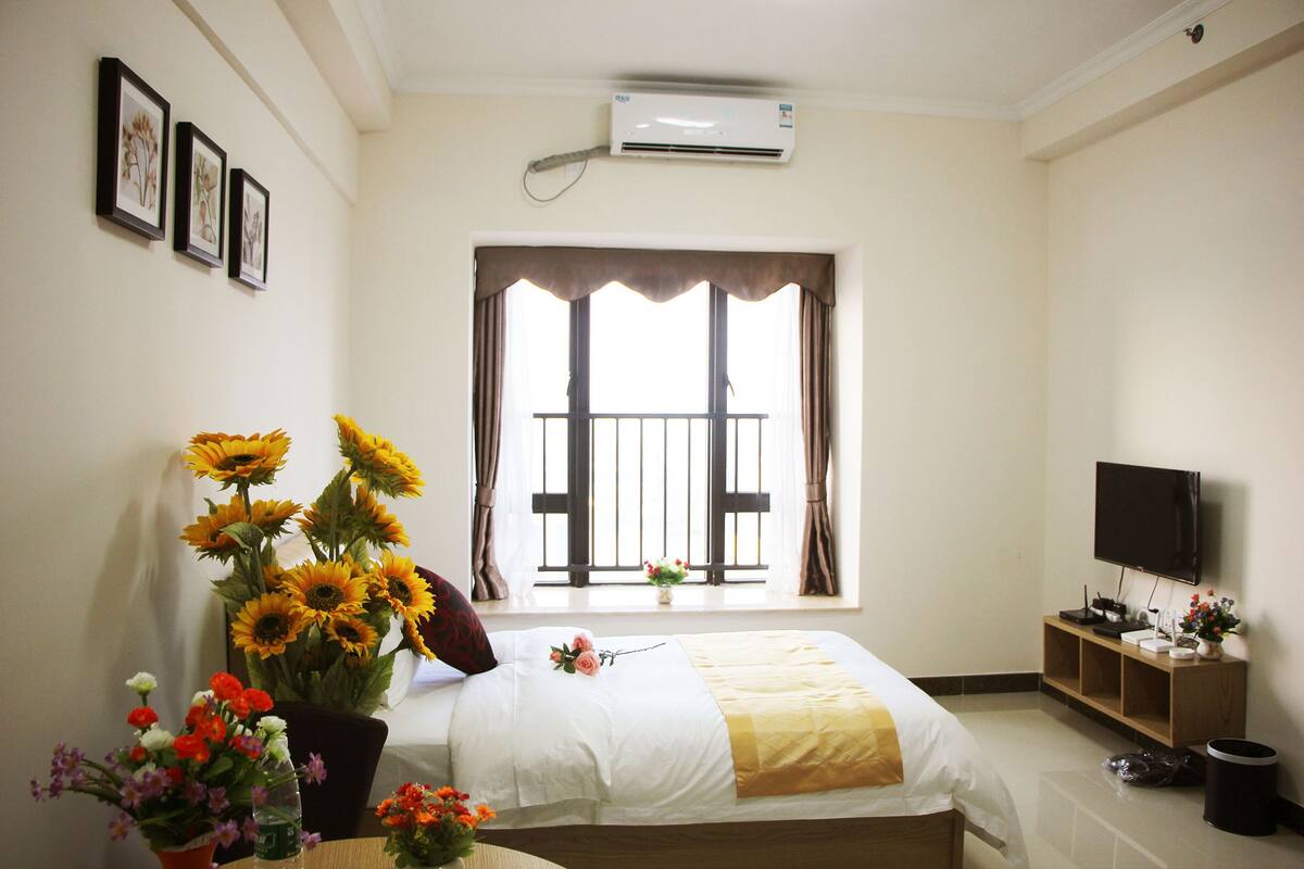 Une chambre d’hôtel avec un lit, une fenêtre avec des rideaux, un climatiseur fixé au mur, une télévision et une petite table agrémentée de fleurs.