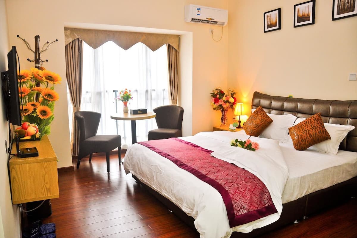 Une chambre d’hôtel avec un lit, un bureau avec un ordinateur, une chaise, une table avec des fleurs et une fenêtre avec des rideaux.