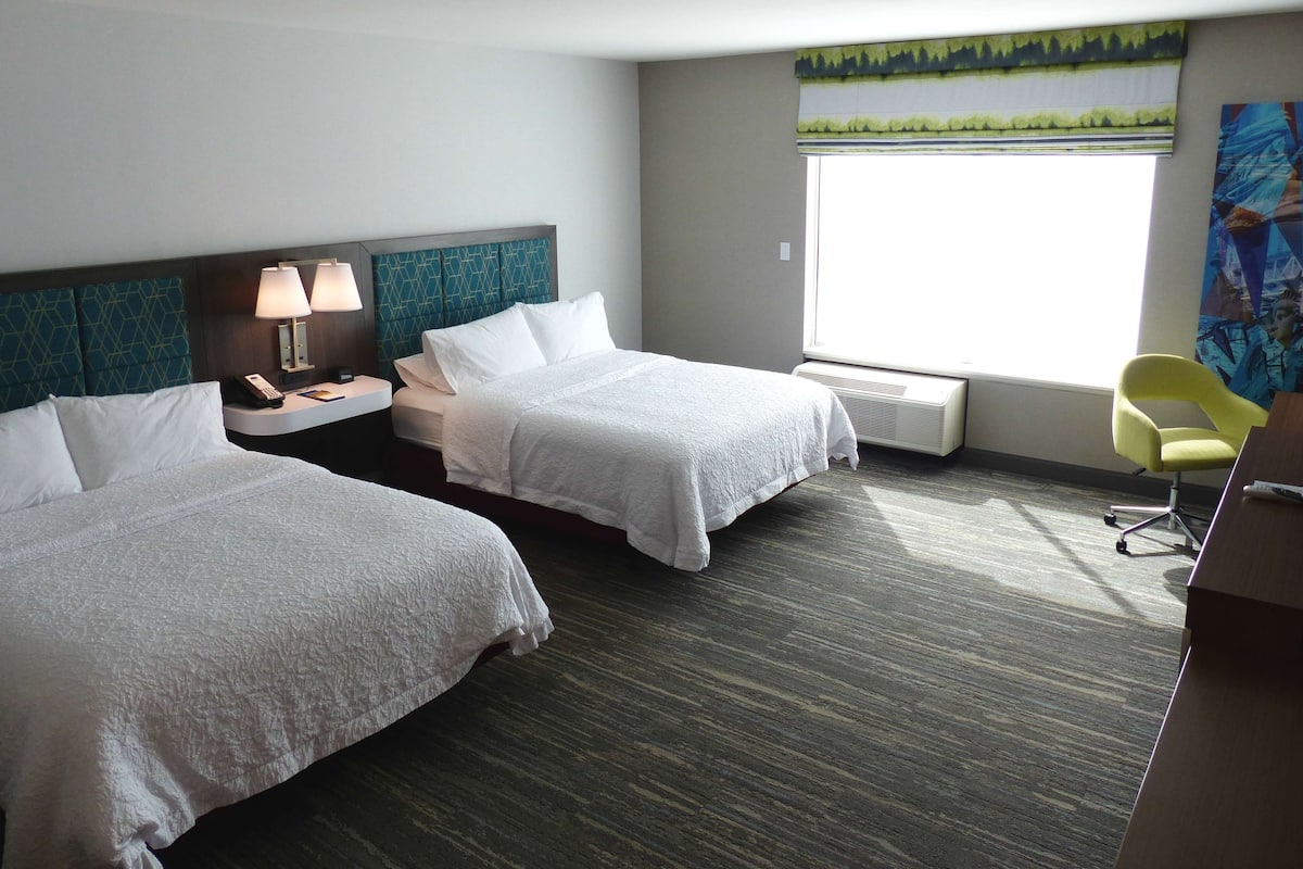 A hotel room with two beds, a desk, a chair, a window with a view, and a painting on the wall.