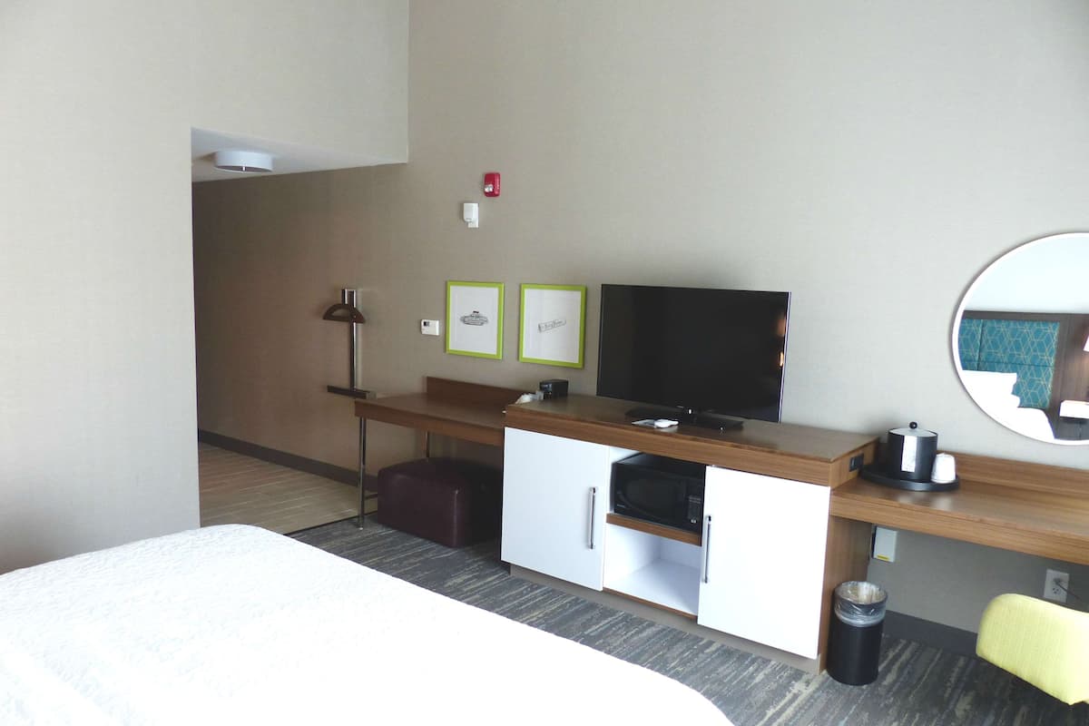 A hotel room with a bed, a desk with a flat-screen TV, a microwave, a trash can, and a chair.