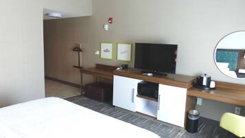 A hotel room with a bed, a desk with a flat-screen TV, a microwave, a trash can, and a chair.