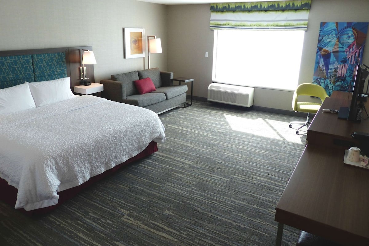 A hotel room with a large bed, a sofa, a desk, and a chair. There is a window with a view of trees and a painting on the wall.