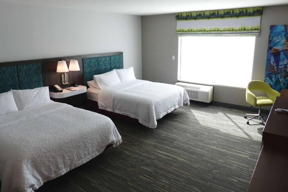 A hotel room with two beds, a desk, a chair, a window with a view, and a painting on the wall.