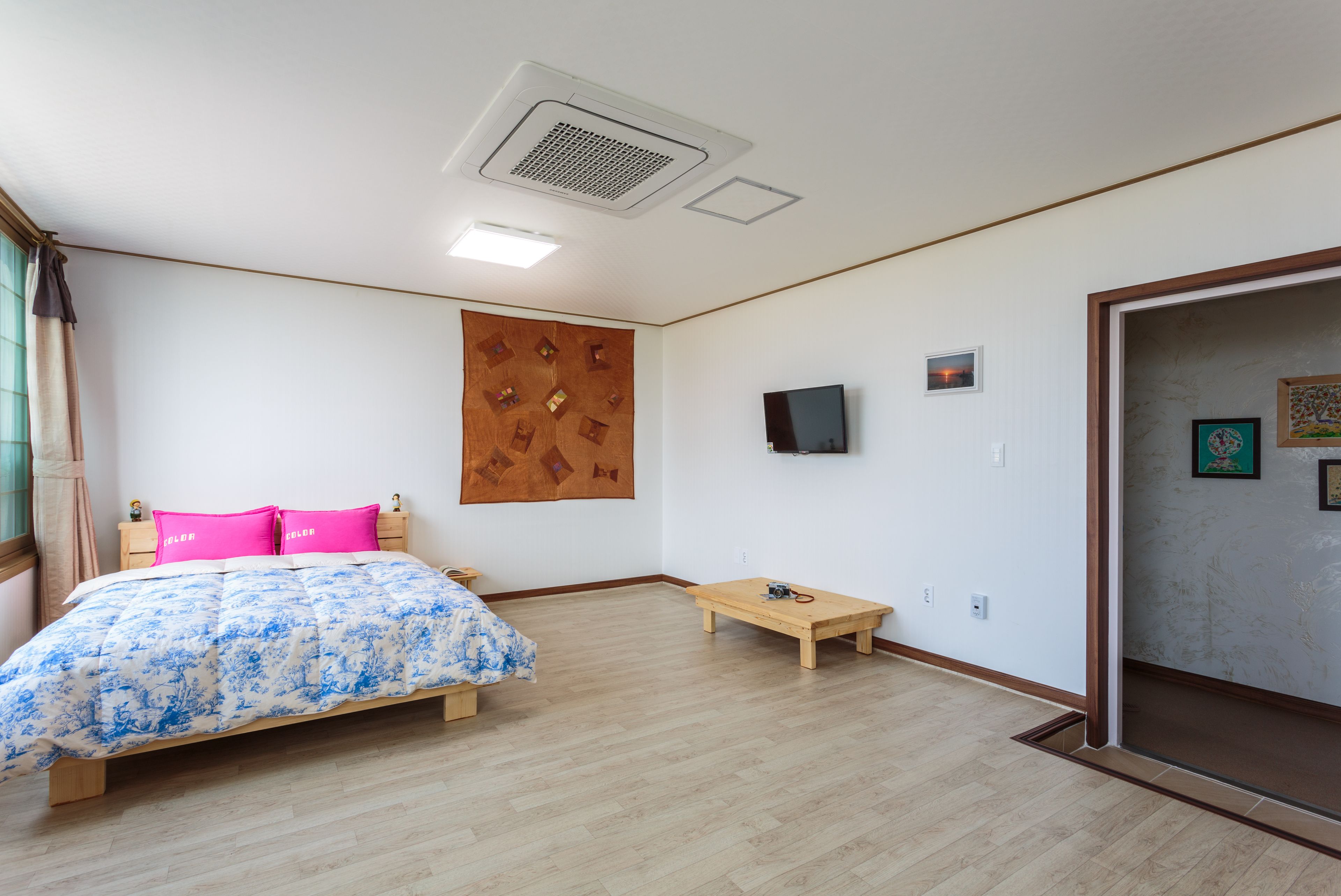 Un dormitorio con cama, cabecera de madera, mesita de noche pequeña, televisor montado en la pared y banco de madera.
