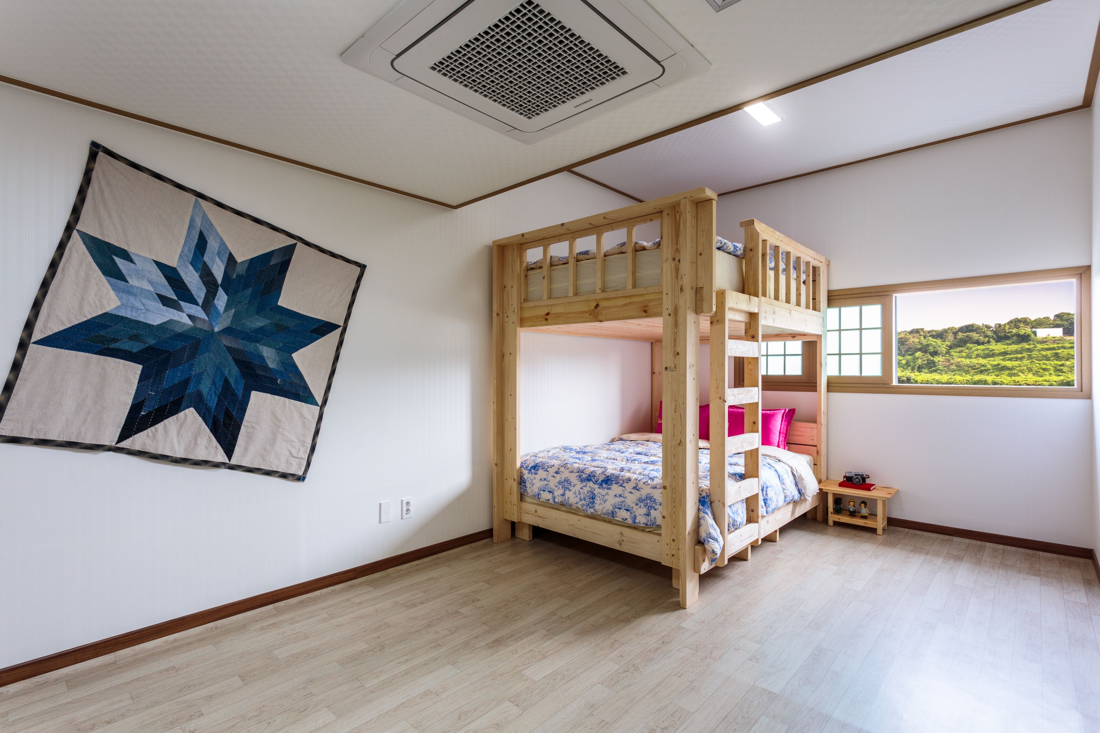 A room with a bunk bed, a star-patterned wall hanging, a small wooden table, and a window with a view of greenery.