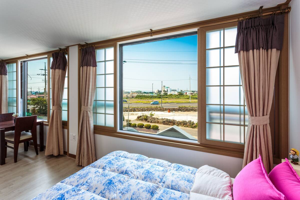 Une chambre avec une grande fenêtre, un lit recouvert d’une literie à motifs, un bureau avec une chaise et une vue sur l’extérieur.
