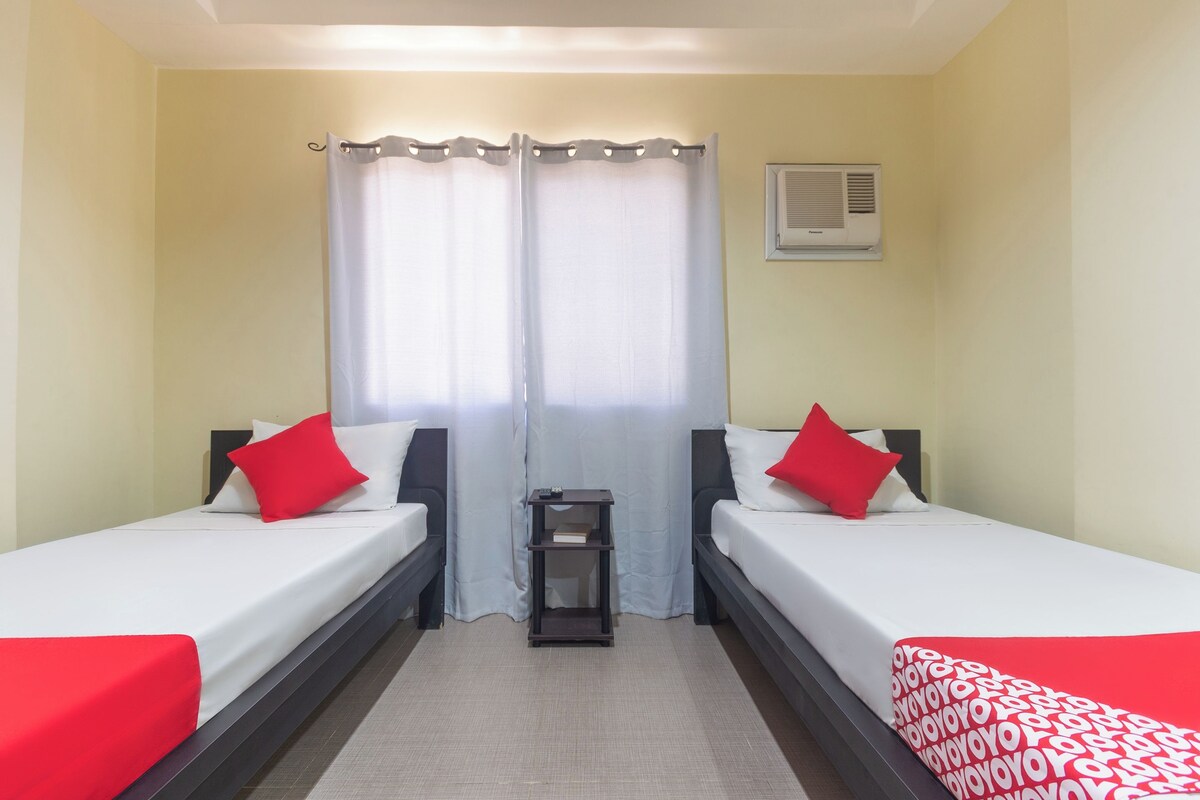A hotel room with two single beds, each with a red pillow, a small bedside table, and a white curtain.