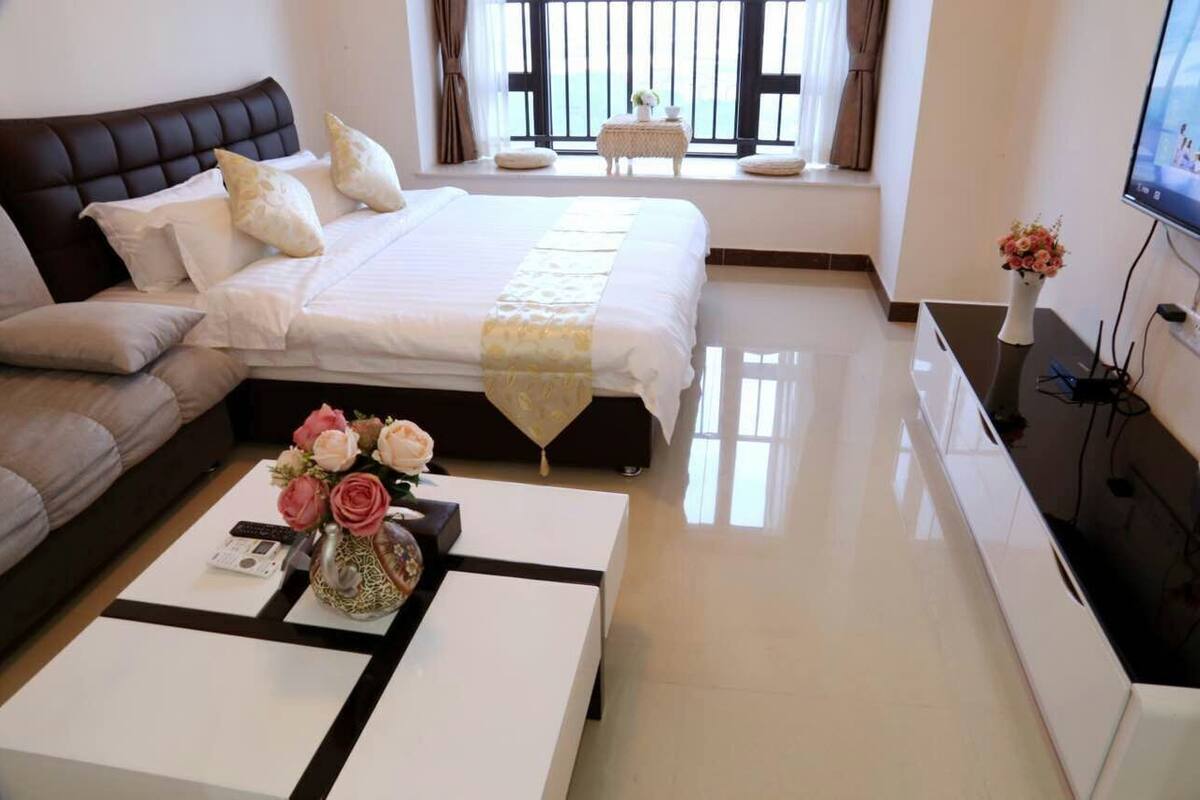 Habitación de hotel con cama, sofá, mesa de centro con un jarrón de flores, televisión y ventana con cortinas.