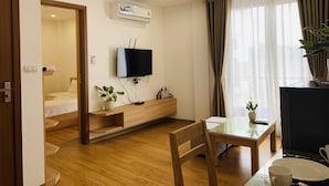 Een moderne hotelkamer met een houten vloer, een glazen eettafel, een aan de muur bevestigde televisie, een zithoekje en een raam met gordijnen.