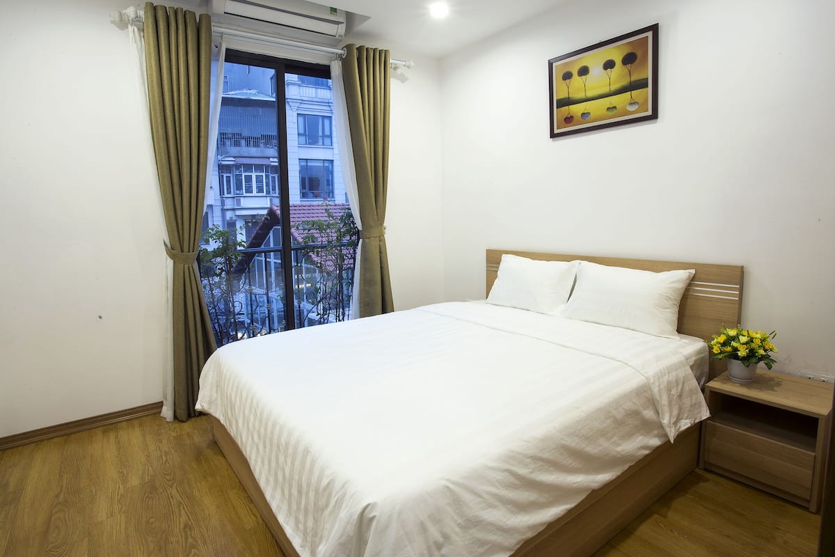 A bedroom with a large bed, a nightstand with a vase of flowers, a window with curtains, and a painting on the wall.