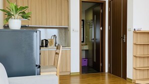 Een moderne woonkamer met een salontafel met glazen blad, een grijze bank, houten vloeren en een keukenhoek met houten keukenkastjes en een potplant.