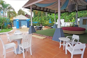 An outdoor patio with white chairs and tables, a covered seating area, and a small blue building in the background.