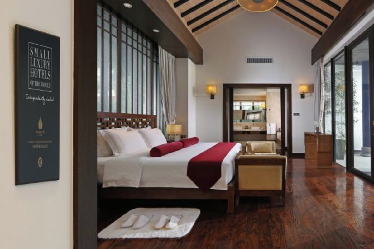 A hotel room with a large bed, a chair, a bedside table, and a view through a large window.