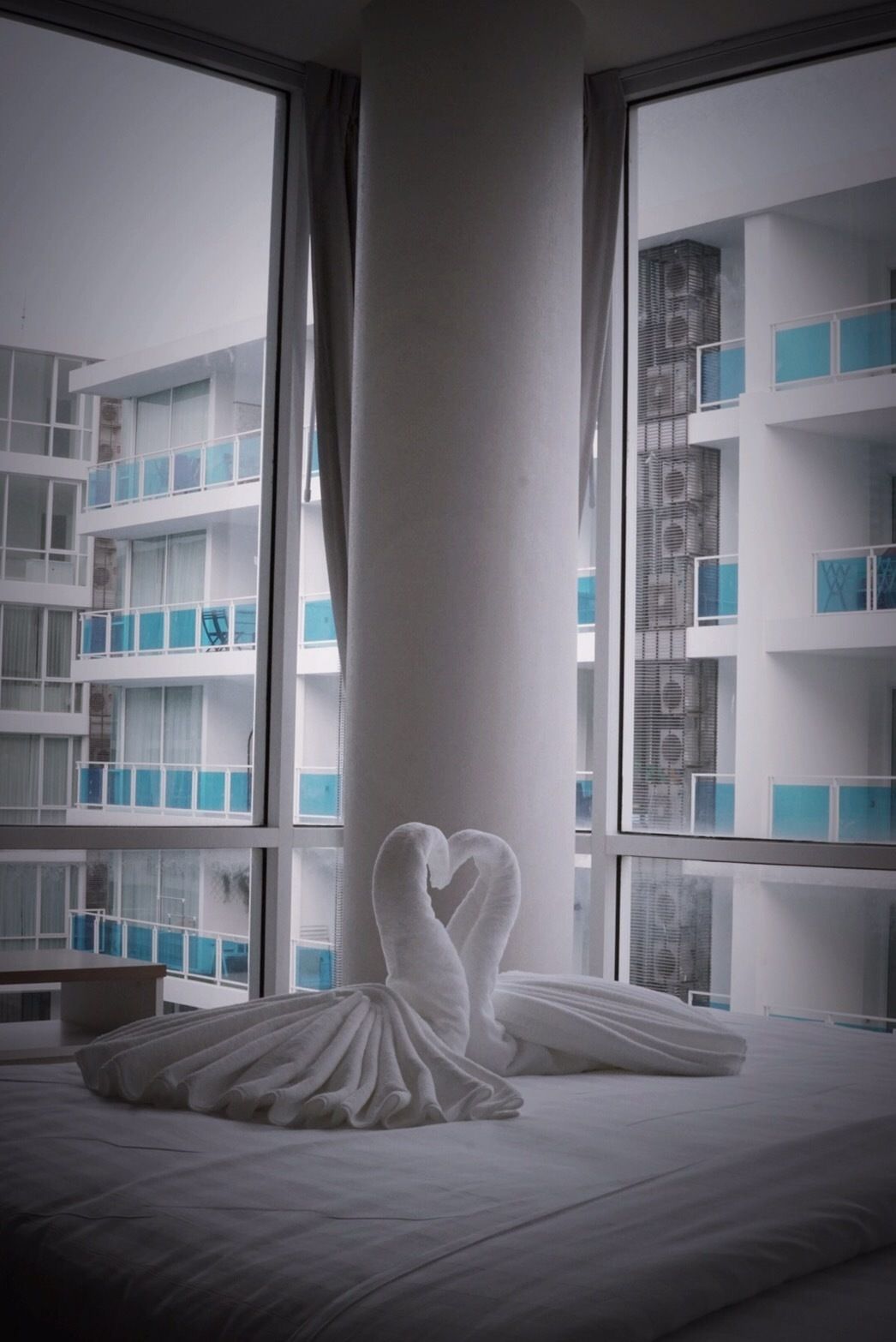 A hotel room with a large window, a bed with a heart-shaped towel, and a view of a multi-story building with balconies.